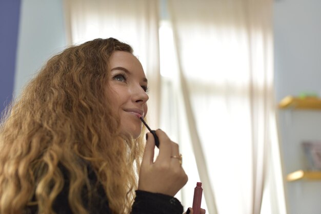 Vista lateral de una mujer hermosa aplicando lápiz labial mientras está sentada en casa