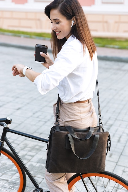 Vista lateral de una mujer joven alegre con ropa de verano caminando por una calle de la ciudad con bicicleta, bebiendo café para llevar