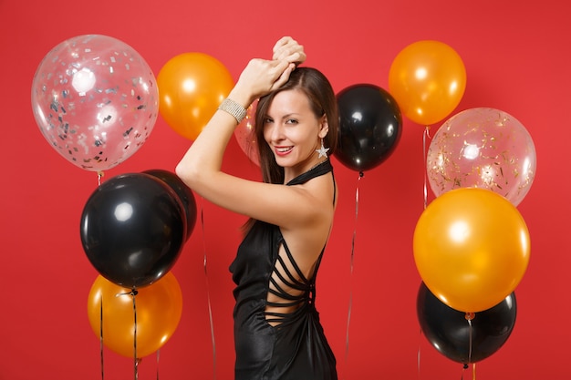 Vista lateral de una mujer joven apasionada en un pequeño vestido negro celebrando, levantando las manos sobre globos de aire de fondo rojo brillante. Día de San Valentín, feliz año nuevo, concepto de fiesta de vacaciones de maqueta de cumpleaños.