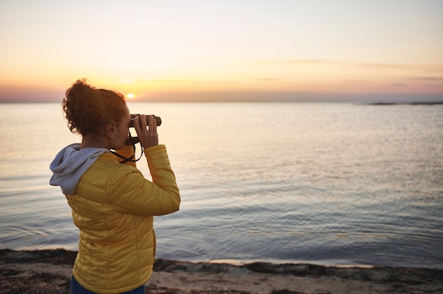 Foto vista lateral de una mujer joven con una chaqueta amarilla mirando a través de binoculares y disfrutando de una hermosa vista del paisaje costero