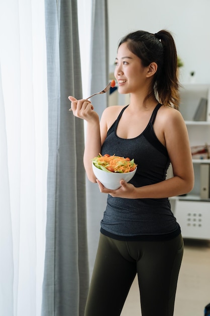 vista lateral mujer joven delgada comiendo ensalada de pie junto a la ventana con sol. chica asiática con ropa de fitness desayunando saludablemente en casa. Mujer en forma con comida vegana después del entrenamiento en el apartamento.