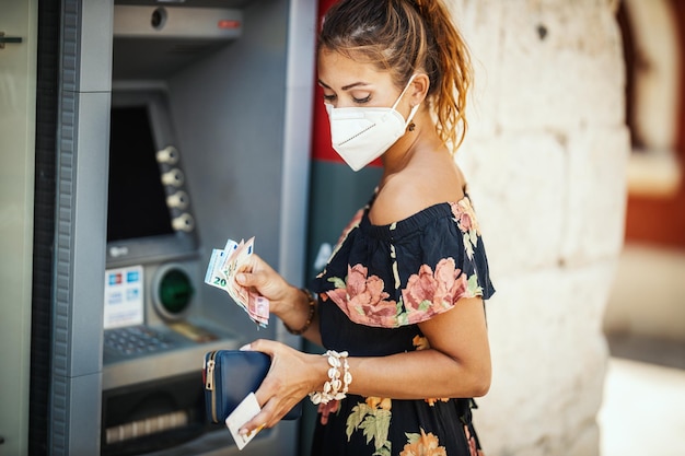 La vista lateral de una mujer joven lleva una máscara protectora N95 mientras saca dinero de un cajero automático durante la pandemia de Covid-19.