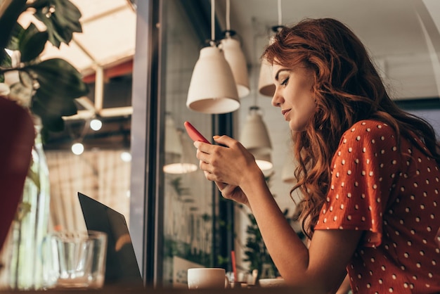 Vista lateral de la mujer joven que usa el teléfono inteligente en la mesa con la computadora portátil en la cafetería