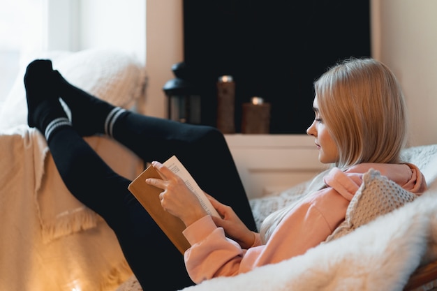 Vista lateral mujer joven relajándose en casa en un cómodo sillón redondo cerca de la ventana. Chica relajante, leyendo y soñando libro en la sala de estar del desván.