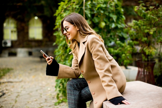 Vista lateral a la mujer joven sentada al aire libre y con teléfono móvil