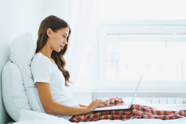 Vista lateral de la mujer joven sentada en la cama en el dormitorio y escribiendo en la computadora portátil