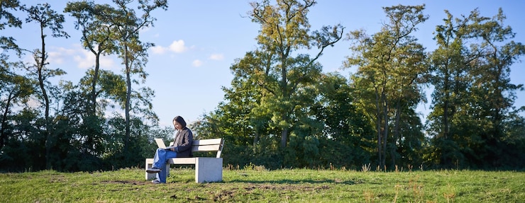 Foto vista lateral de una mujer joven sentada sola en un parque en un banco usando su computadora portátil para estudiar o trabajar de forma remota