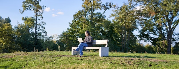 Vista lateral de una mujer joven sentada sola en un parque en un banco usando su computadora portátil para estudiar o trabajar de forma remota