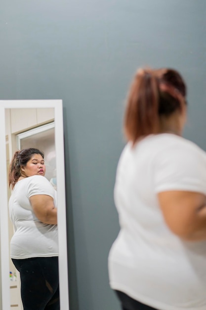 Foto vista lateral de una mujer joven con sobrepeso mirándose en el espejo mientras está de pie en casa