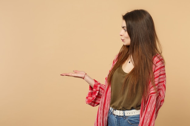 Vista lateral de la mujer joven sorprendida en ropa casual mirando y apuntando con la mano a un lado aislado sobre fondo de pared beige pastel en estudio. Personas sinceras emociones, concepto de estilo de vida. Simulacros de espacio de copia.