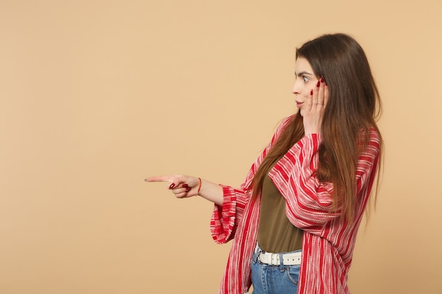 Vista lateral de la mujer joven sorprendida en ropa casual poniendo la mano en la mejilla apuntando con el dedo índice a un lado aislado sobre fondo beige pastel. Personas sinceras emociones, concepto de estilo de vida. Simulacros de espacio de copia.