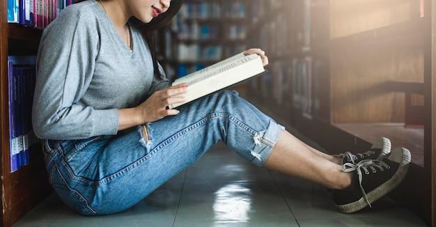 Foto vista lateral de una mujer leyendo un libro mientras está sentada en el piso de azulejos en la biblioteca