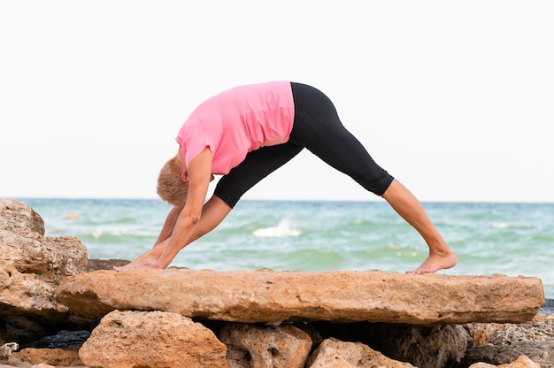 Vista lateral de la mujer mayor practicando yoga en la playa.