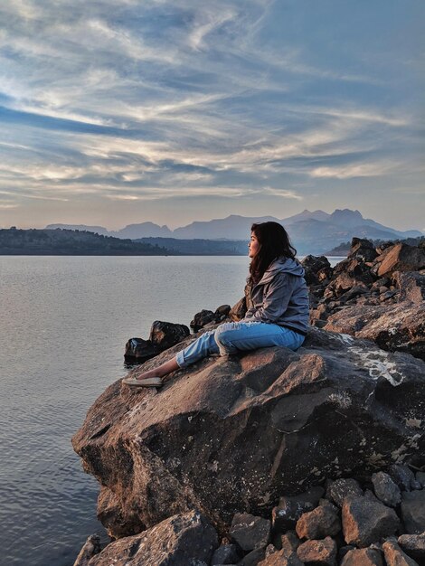 Vista lateral de una mujer mirando al mar mientras está sentada en rocas contra el cielo durante la puesta de sol