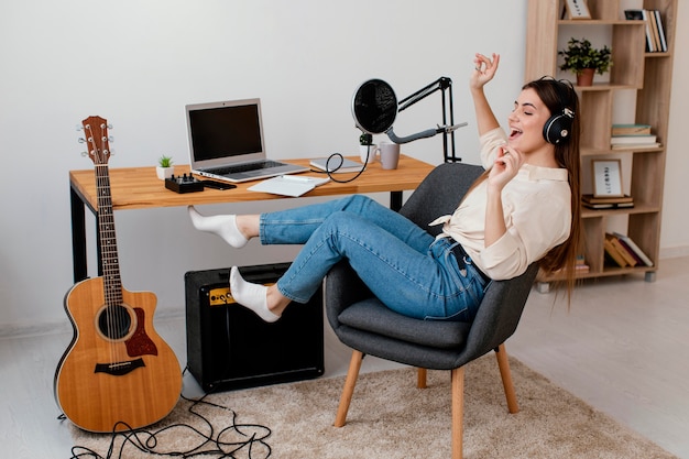 Vista lateral de la mujer músico en casa cantando con auriculares junto a la guitarra acústica