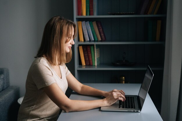 Vista lateral de una mujer de negocios enfocada que trabaja en una computadora portátil desde la oficina en casa sentada en el escritorio en una habitación oscura