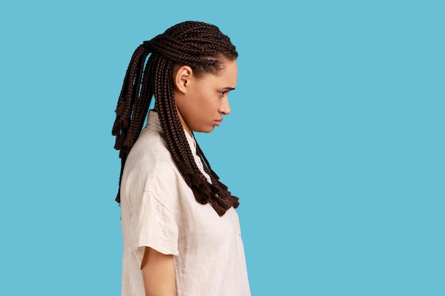 Vista lateral de una mujer ofendida con dreadlocks mirando con el ceño fruncido, espera explicaciones o disculpas, irritada por algo injusto. Disparo de estudio interior aislado sobre fondo azul.