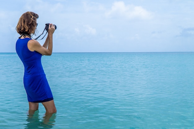 Foto vista lateral de una mujer de pie sobre el agua y tomar fotografías del mar caribe.