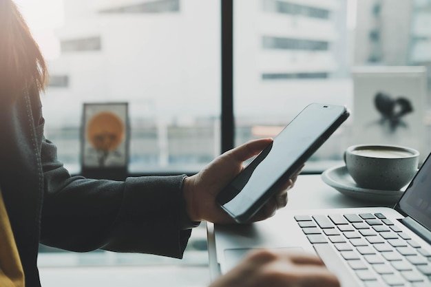 Vista lateral de la mujer que usa un teléfono inteligente negro y una computadora portátil en la mano y lo señala con el dedo mientras está sentada en el escritorio de trabajo