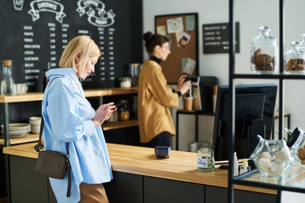 Vista lateral de la mujer rubia madura enviando mensajes de texto en el teléfono inteligente en el café