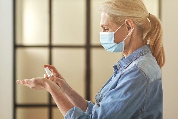 Vista lateral de la mujer rubia madura con mascarilla protectora aplicando desinfectante de manos antibacteriano