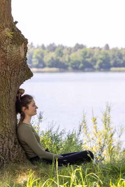 Foto vista lateral de una mujer sentada contra un árbol junto al lago