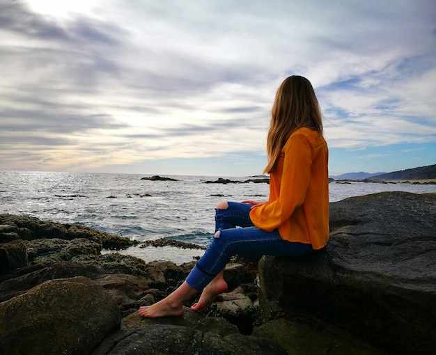 Vista lateral de una mujer sentada en una roca en la playa contra un cielo nublado