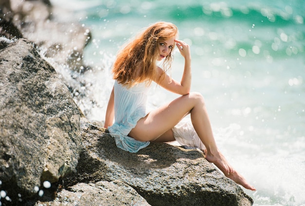 Vista lateral de la mujer serena sentada en la playa rocosa de arena contra las olas azules atractiva mujer joven wea