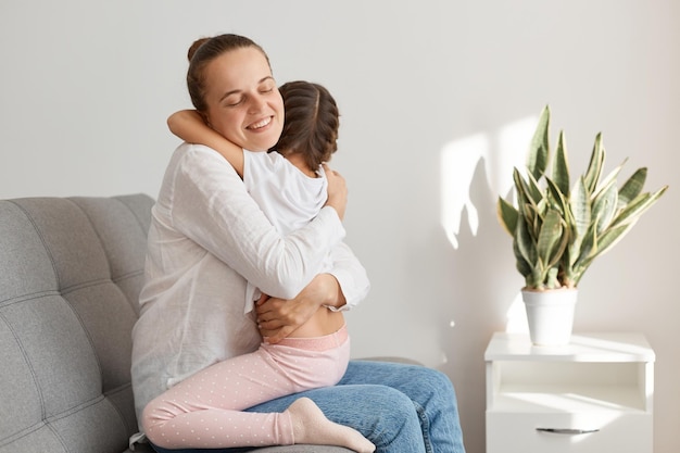 Vista lateral de una mujer sonriente con moño con camisa blanca y jeans, abrazando a una niña que está sentada de rodillas, familia posando en casa mientras se sienta en un cómodo sofá.