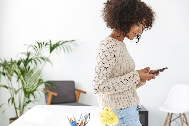 Vista lateral de la mujer sonriente vestida con ropa casual con teléfono inteligente mientras está de pie cerca de la mesa en la oficina