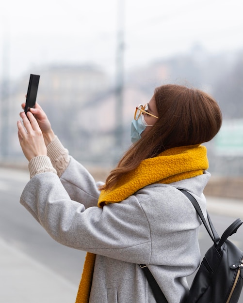 Foto vista lateral de la mujer tomando fotografías con el teléfono inteligente mientras usa una máscara médica