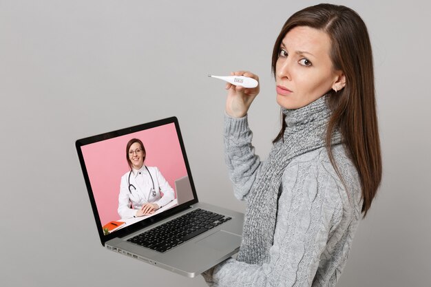 Vista lateral de la mujer trastornada en la bufanda del suéter que sostiene la computadora portátil del pc del termómetro con la pantalla vacía en blanco aislada en fondo gris. Consulta de tratamiento en línea de estilo de vida saludable concepto de estación fría.