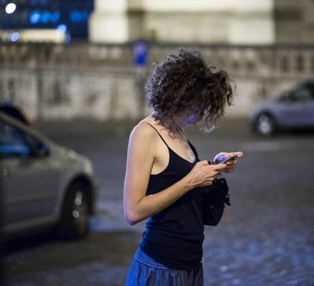 Vista lateral de una mujer usando el teléfono móvil mientras está de pie en la calle por la noche