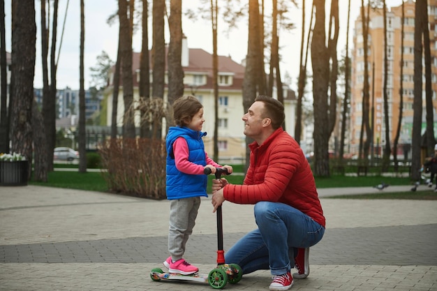 Vista lateral de una niña adorable en una patineta sonriendo alegremente mientras mira a su padre de pie cara a cara con él en un parque de la ciudad en un hermoso día soleado de otoño