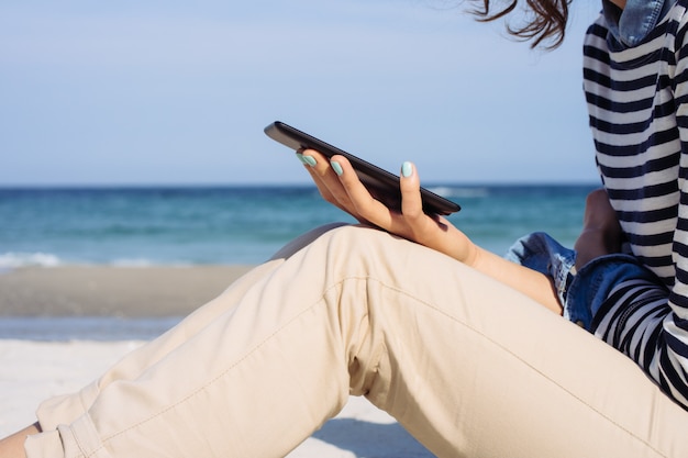 Vista lateral de una niña con una camiseta a rayas y pantalones beige en la playa que está leyendo un libro