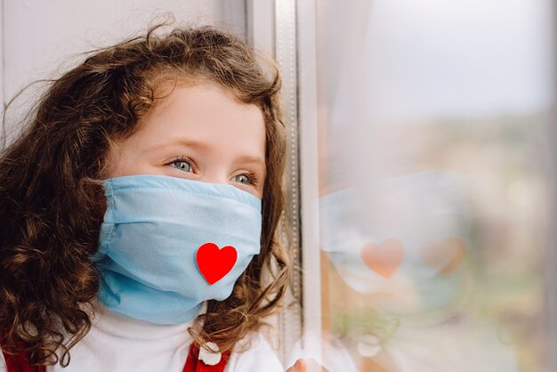 Vista lateral de la niña en edad preescolar en mascarilla protectora con un corazón rojo, sentado en el alféizar de la ventana en casa, mirando fuera de la ventana borrosa. Concepto gracias a todos los empleados esenciales durante la pandemia de Covid-19