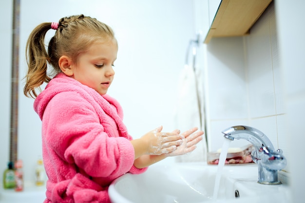 Vista lateral de la niña linda con cola de caballo en bata de baño rosa que se lava las manos.