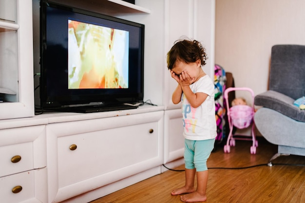 Vista lateral de una niña linda viendo la televisión con juguetes en el piso en casa La niña pequeña está llorando cerca de ver la televisión