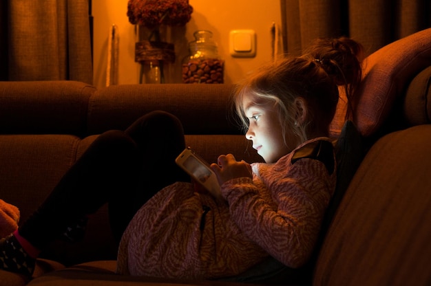 Vista lateral de una niña usando el teléfono móvil mientras está sentada en el sofá en casa