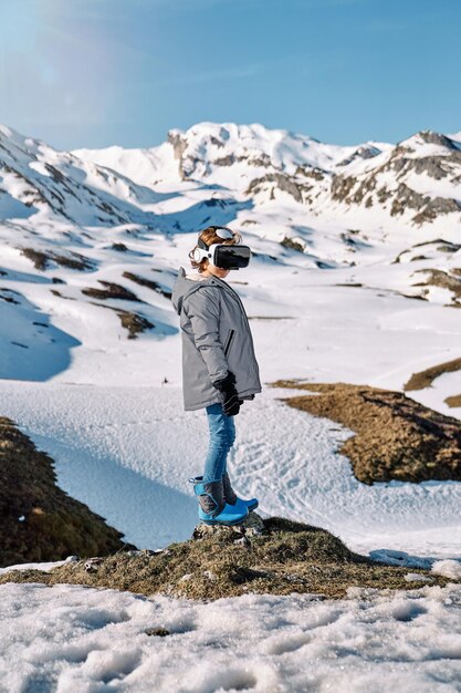 Vista lateral de un niño anónimo con ropa abrigada y gafas VR parados en un terreno rocoso cerca de montañas nevadas mientras explora el mundo virtual