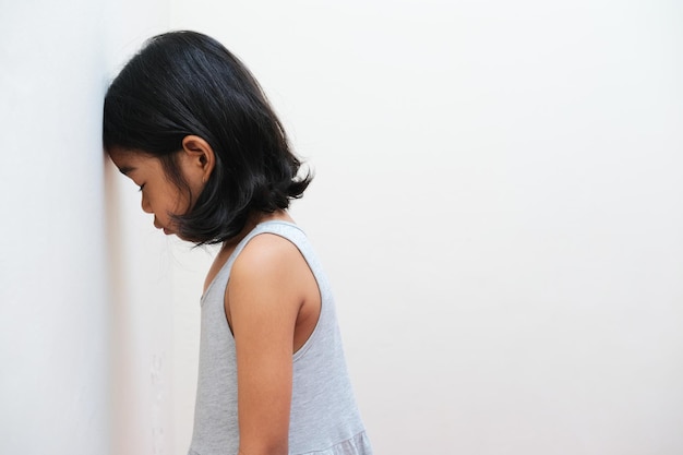 Vista lateral de un niño asiático que muestra una expresión triste con la cabeza en la pared