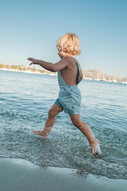 Vista lateral de un niño descalzo anónimo de cuerpo completo disfrutando y jugando en el mar ondulante bajo un cielo azul brillante