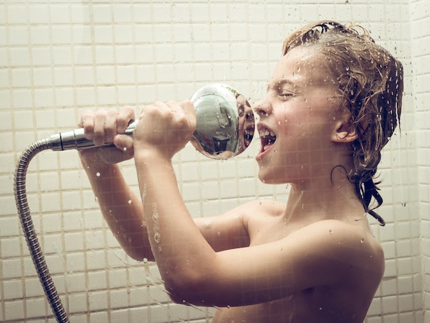 Vista lateral de un niño desnudo sosteniendo el cabezal de la ducha y fingiendo cantar en el baño bajo el agua corriente