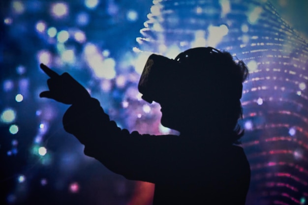 Foto vista lateral de un niño irreconocible con modernas gafas vr tocando una pantalla imaginaria mientras explora la realidad virtual en una habitación oscura con una brillante iluminación de neón