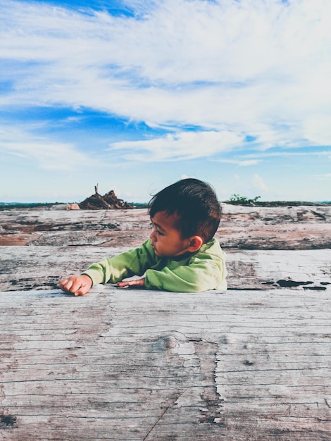 Foto vista lateral de un niño sentado junto al tronco de un árbol contra el cielo