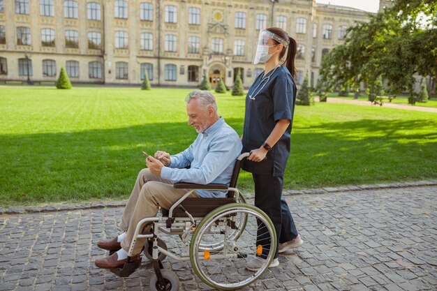 Vista lateral del paciente de recuperación del hombre mayor en silla de ruedas con smartphone en una caminata con la enfermera