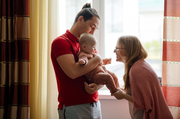 Vista lateral del padre sosteniendo al bebé mientras la madre habla con el bebé contra la ventana