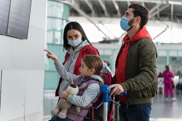 Vista lateral de los padres y su hija con máscaras protectoras frente al tablero de horarios mientras esperan el vuelo en el aeropuerto. Viajar durante el concepto de pandemia