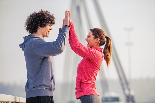 Foto vista lateral de la pareja caucásica deportiva dando cinco altos entre sí mientras está de pie en el puente en clima frío. concepto de gimnasio al aire libre.