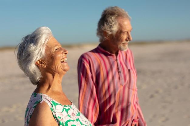 Vista lateral de una pareja caucásica mayor en la playa bajo el sol, cogidos de la mano y mirando al mar, la mujer riendo, con el cielo azul de fondo